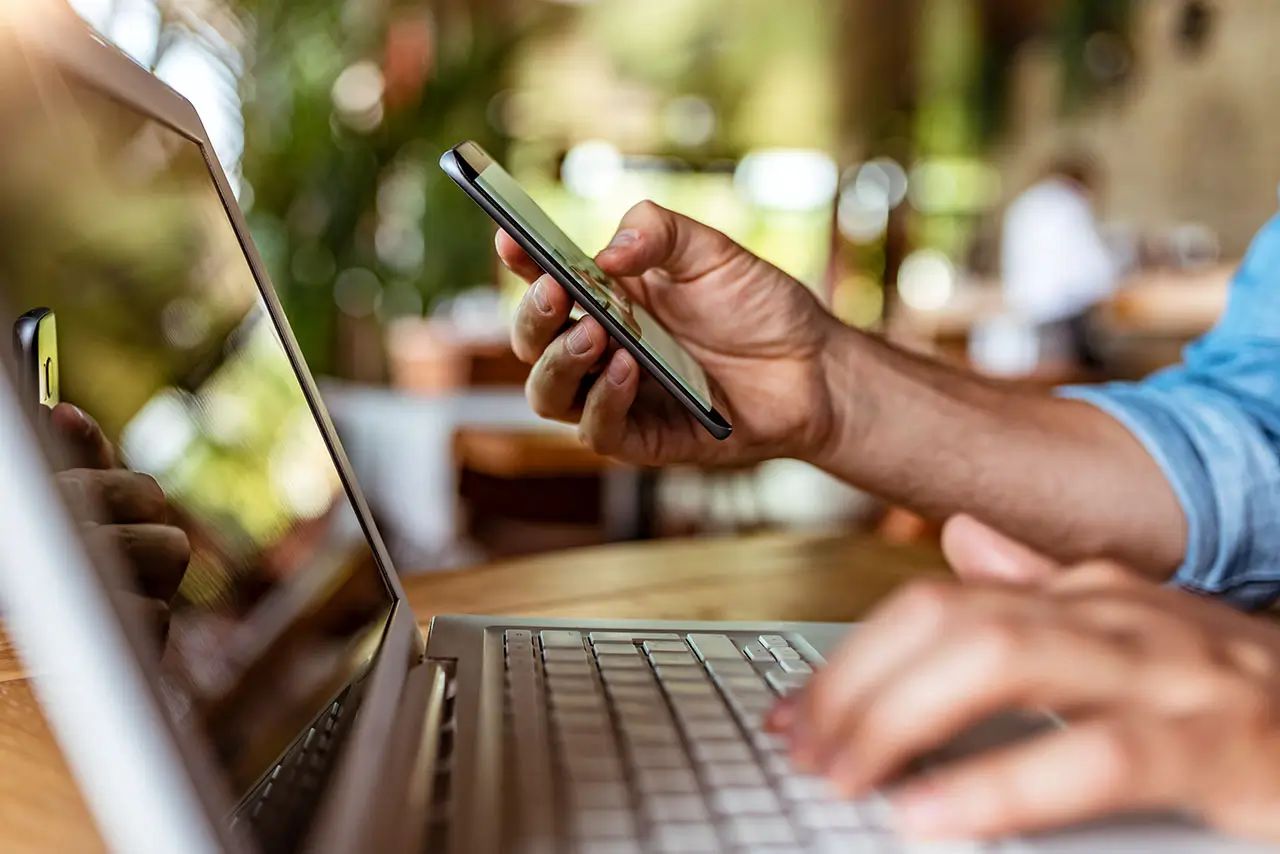 A closeup view of someone working on their laptop while looking at their cellphone.